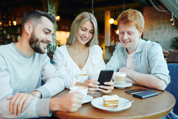 Pretty blonde girl with smartphone showing two happy guys her new photos in cafe - Stock Photo - Images