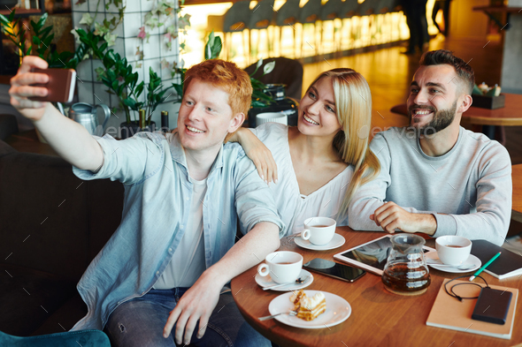 Three happy young affectionate friends making selfie in cozy cafe by cup of tea