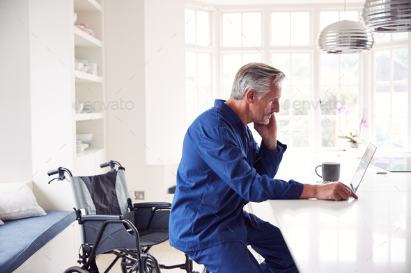 Mature Disabled Man In Wheelchair At Home Using Laptop On Kitchen Counter