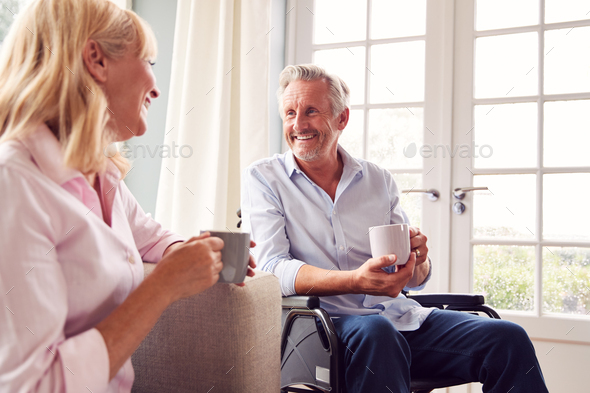 Mature Couple With Man In Wheelchair Sitting In Lounge At Home Talking Together