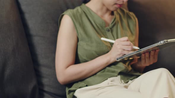 Asian woman reading news on her digital tablet while sitting on the sofa in the living room.