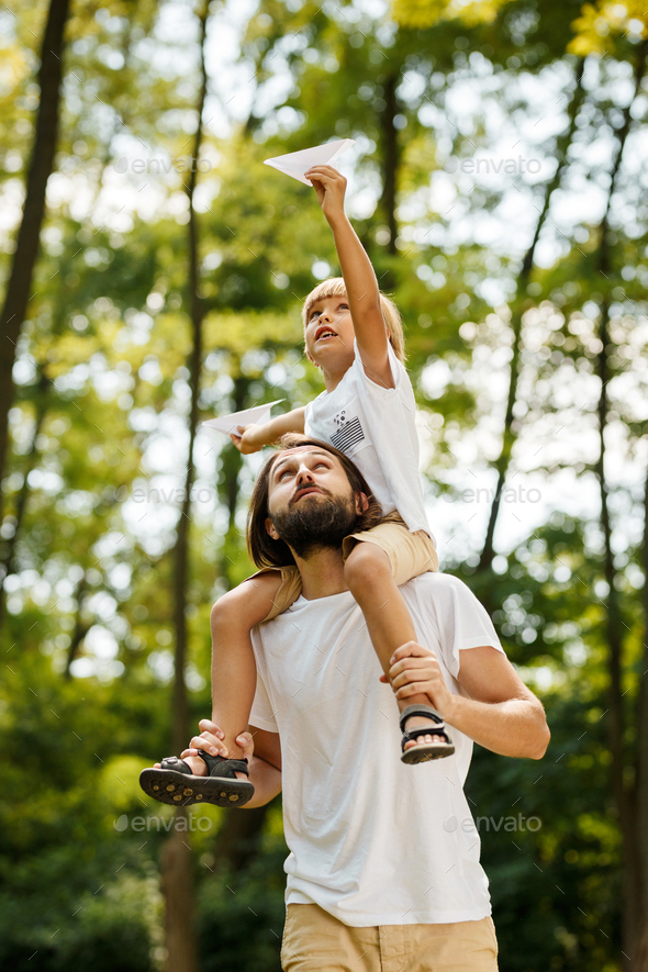 Walking in the forest. Blond boy dressed in a white t-shirt sitting on the shoulders of his bearded - Stock Photo - Images