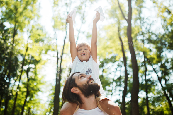 Handsome father and his little son walking in the forest. Boy is sitting on the shoulders of his - Stock Photo - Images