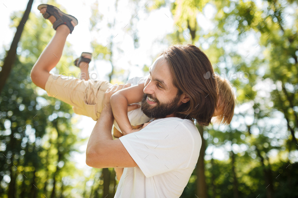 Dark-haired father with beard dressed in the white t-shirt is holding in the arms his blond son and - Stock Photo - Images