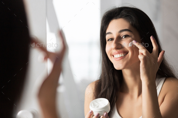 Cheerful Girl Applying Face Cream Looking In Mirror Indoor