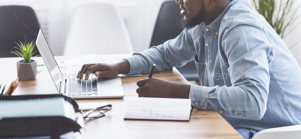 Black trainee taking notes while using laptop in office