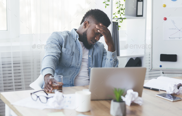 Depressed black businessman drinking alcohol at workplace in office