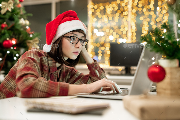 Serious young businesswoman in Santa cap sitting by desk in front of laptop