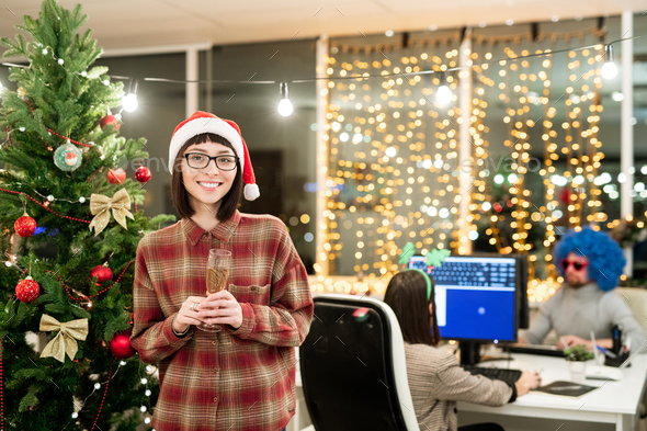 Cheerful female office manager with flute of champagne standing by xmas tree