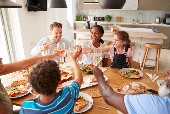 Multi-Generation Mixed Race Family Making A Toast Before Eating Meal Around Table At Home Together