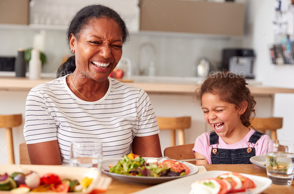 Grandmother And Granddaughter Laughing As They Eat Meal At Table