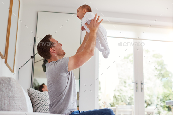 Loving Father Lifting 3 Month Old Baby Daughter In The Air In Lounge At Home