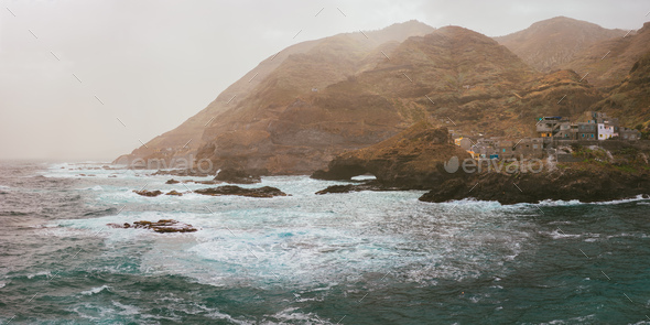 Bluff volcanic coast line. Sunlight penetrates dust and fall over sea foam and steep slopes - Stock Photo - Images