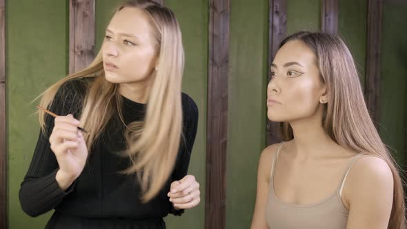 Attractive Caucasian Makeup Artist Doing Makeup for Asian Female in Beauty Salon alt