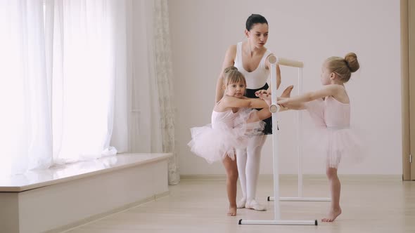 Two Little Girls Practicing Choreographic Elements on Ballet Barre with Help of Teacher in Dance alt
