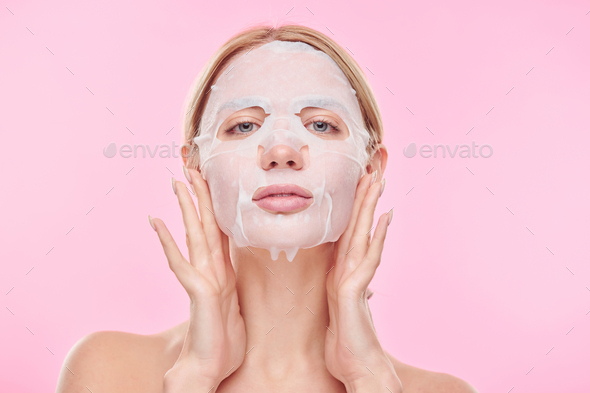 Young woman putting refreshening textile mask on face while taking care of skin