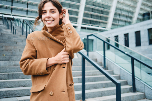 Young positive woman fastening up coat from cold happily looking in camera outdoor