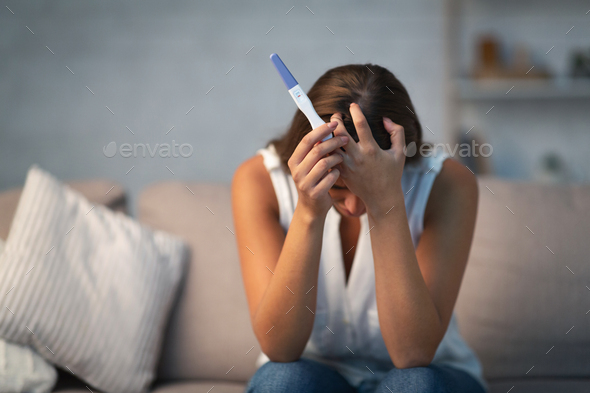 Desperate woman holding pregnancy test sitting on sofa at home