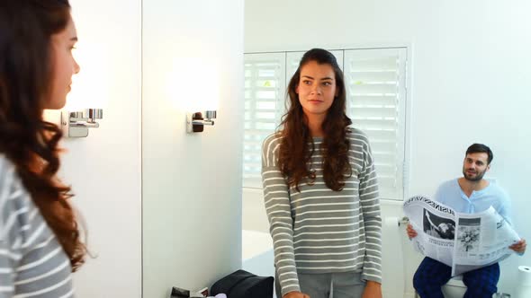 Woman looking at mirror while man reading newspaper in background alt