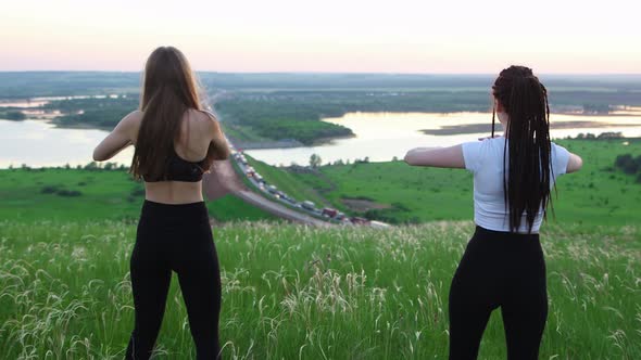 Two Young Women Doing Warm Up Exercises on the Green Field While Late Sunset alt