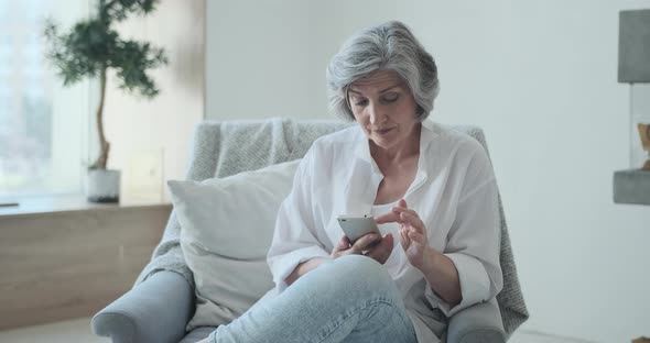 Older Middle Aged Female Looking at Smartphone Screen Watching Funny Photo or Video alt