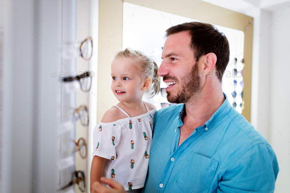 Health care, eyesight and vision concept. Little girl choosing glasses with father at optics store