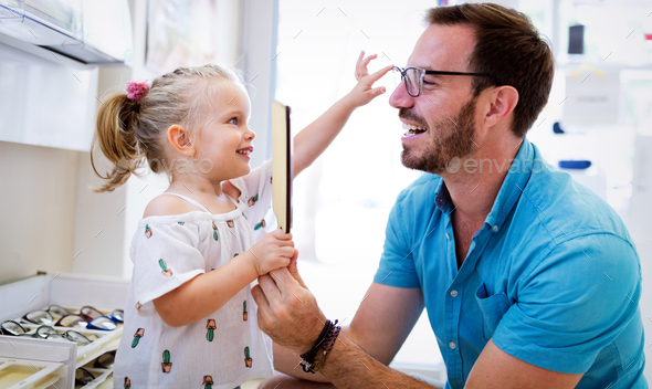 Health care, eyesight and vision concept. Little girl choosing glasses with father at optics store