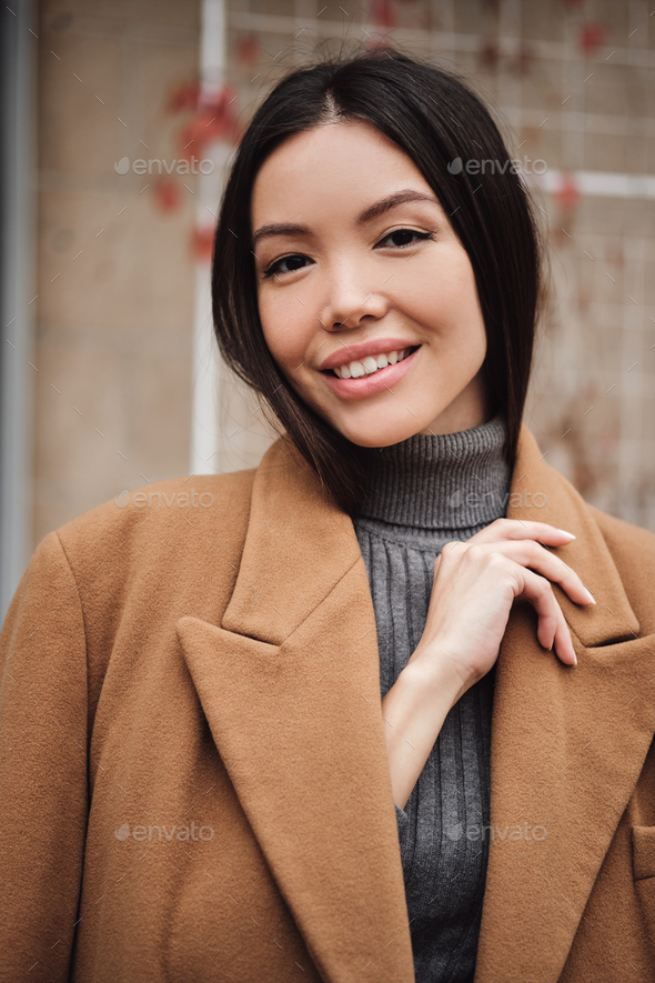 Portrait of gorgeous Asian girl in coat happily looking in camera outdoor