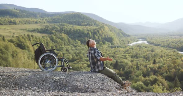 Likable Happy Smiling Young Disabled Man Relaxing on the Top of the Hill and Stretching His Hands in alt