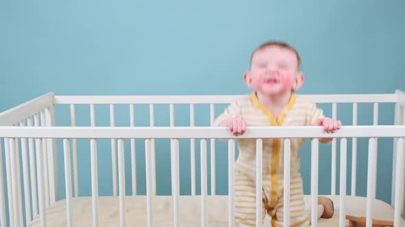 Cry infant baby boy stands in the crib, studio blue background. Sad child in yellow pajamas alt