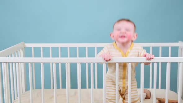 Cry infant baby boy stands in the crib, studio blue background. Sad child in yellow pajamas