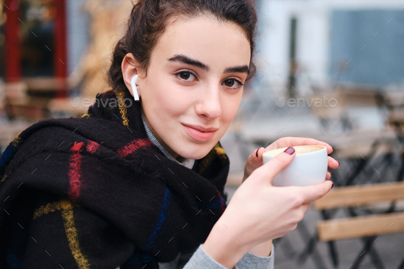Pretty girl with wireless earphones happily looking in camera drinking coffee in cafe outdoor