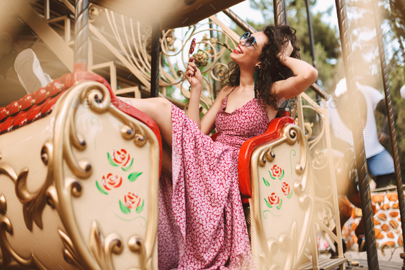 Pretty girl in sunglasses and dress holding lolly pop happily riding on carousel in amusement park - Stock Photo - Images