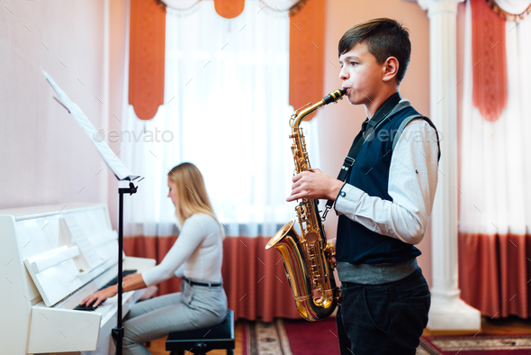Boy student learns to play the saxophone in a music lesson to accompaniment of the piano.