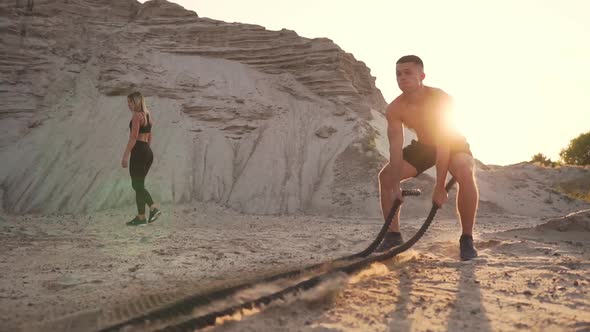 Athletic Man on Exercise Around the Sand Hills at Sunset Hits the Rope on the Ground alt