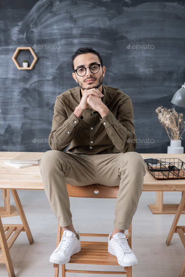 Young restful businessman in casualwear looking at you while sitting on table - Stock Photo - Images