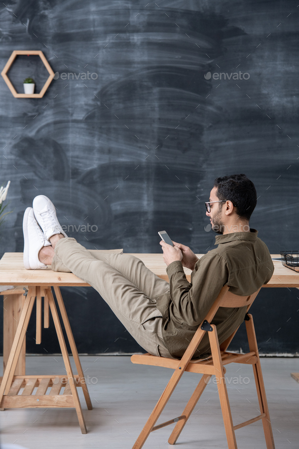 Young casual employee keeping legs on table while scrolling in smartphone - Stock Photo - Images