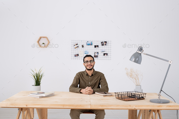 Young successful businessman in casualwear sitting by wooden table in office