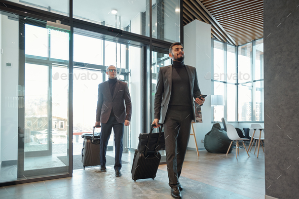 Two elegant business travelers pulling suitcases while entering hotel lounge - Stock Photo - Images