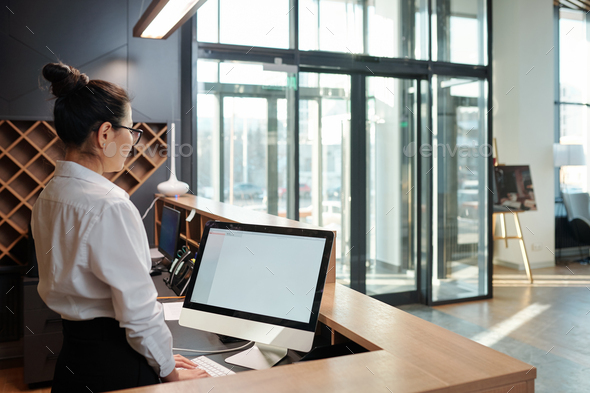 Young female receptionist standing in front of computer screen by counter - Stock Photo - Images