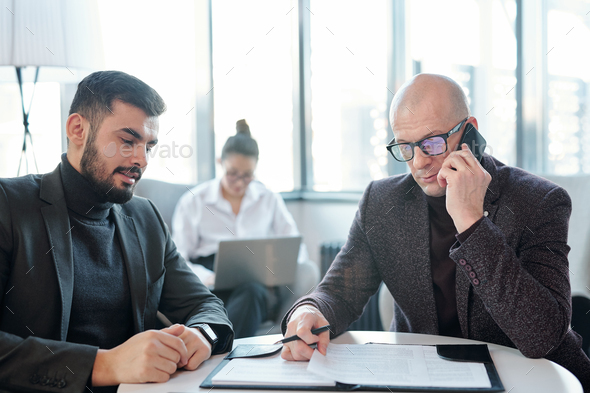 Busy broker in formalwear looking through papers and consulting client by phone - Stock Photo - Images
