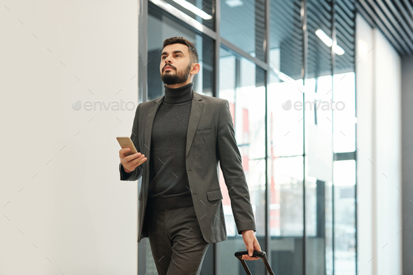 Young businessman in formalwear pulling suitcase while moving towards exit