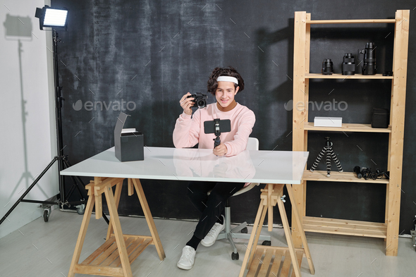 Male vlogger sitting by desk and showing photocamera while shooting himself