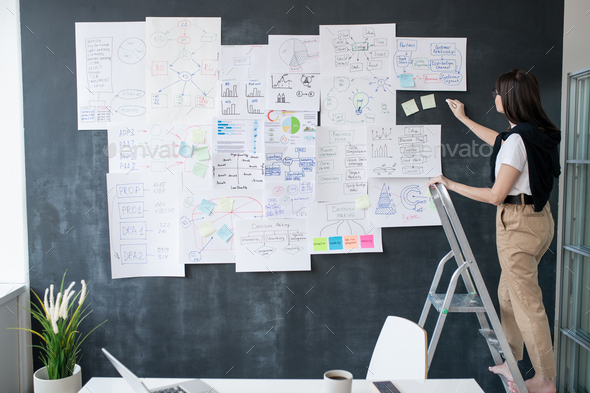Young woman with piece of chalk standing on step ladder in front of blackboard - Stock Photo - Images