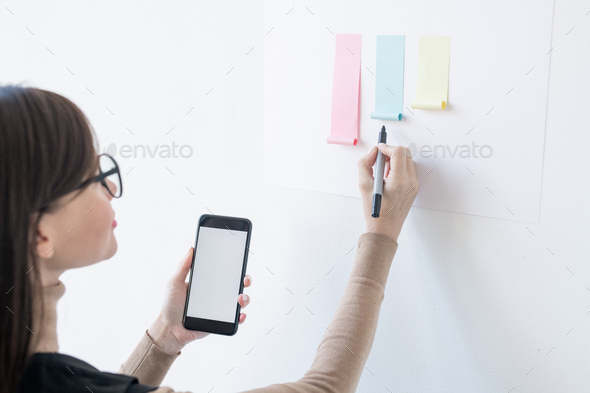 Young female broker with smartphone and highlighter looking at paper chart - Stock Photo - Images