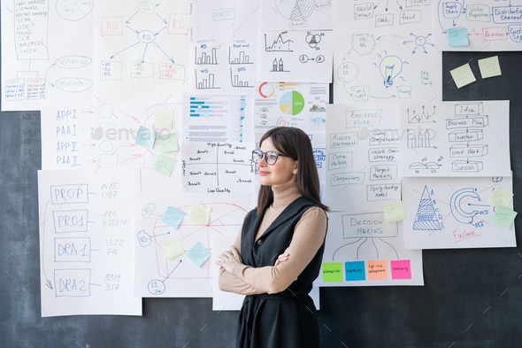 Young successful female economist with crossed arms standing by blackboard - Stock Photo - Images