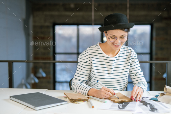 Happy young creative female sitting by desk in studio and drawing fashion sketch - Stock Photo - Images