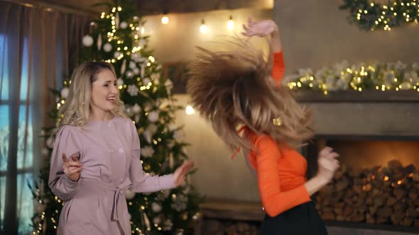 Two Blondes in Festive Clothes Dance Against the Background of a Christmas Tree and Lights alt