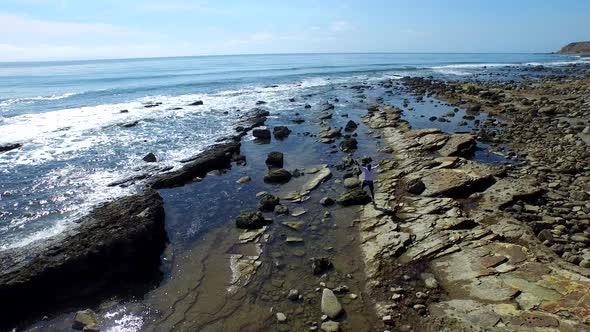 Aerial shot of young man running on a scenic rocky beach coastline. alt