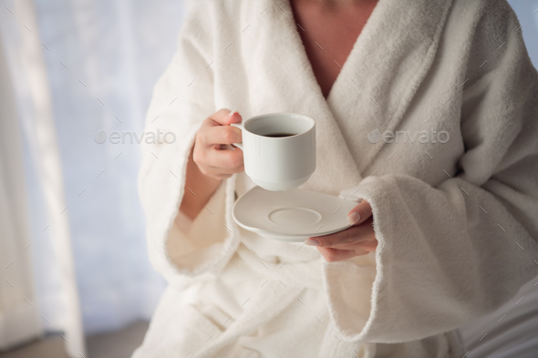 Attractive young girl in bathrobe holding a cup, looking at window and smiling while sitting on a - Stock Photo - Images